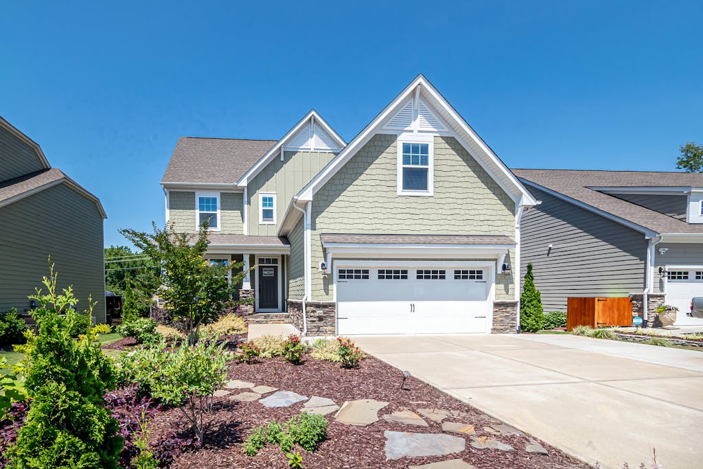 Charming suburban house with green facade, garage, and landscaped yard on a clear day.